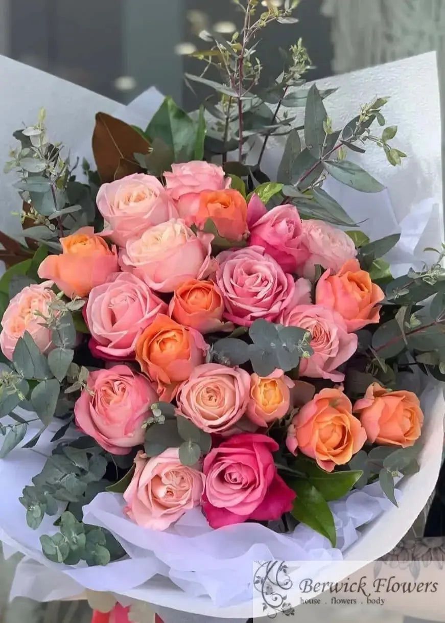 Bouquet of pink, orange, and red roses with greenery on a white surface.