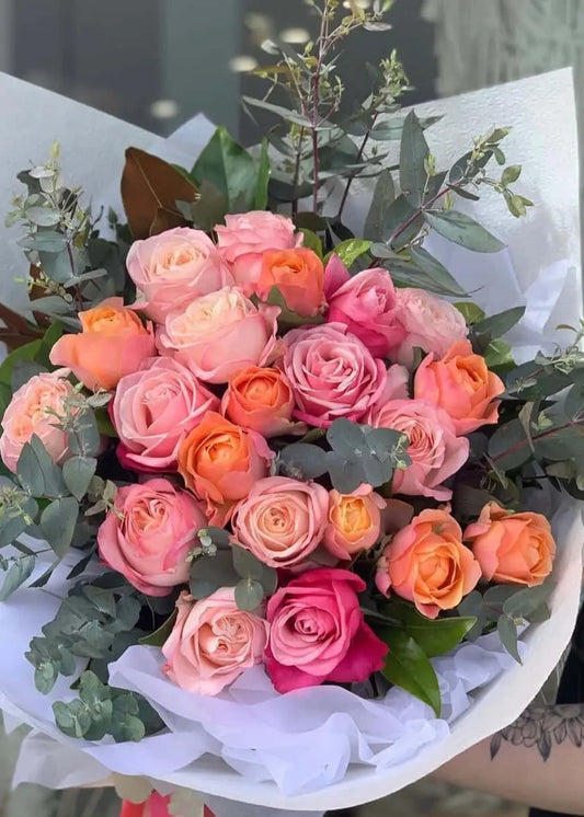 Bouquet of pink, orange, and red roses with greenery on a white surface.