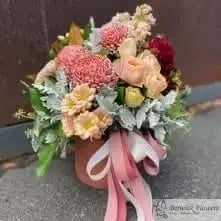 A small floral arrangement in a terracotta-colored pot, featuring pink, peach, yellow, and red flowers with green foliage and a pink ribbon, placed on a gray surface.