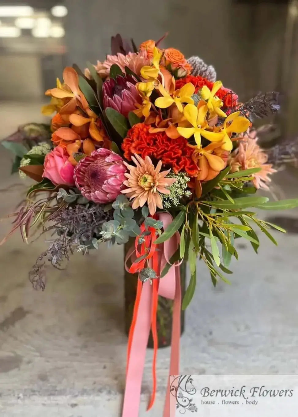 A vibrant autumn floral bouquet featuring orange, pink, yellow, and red flowers with green foliage, tied with a pink ribbon, set against a blurred concrete background.