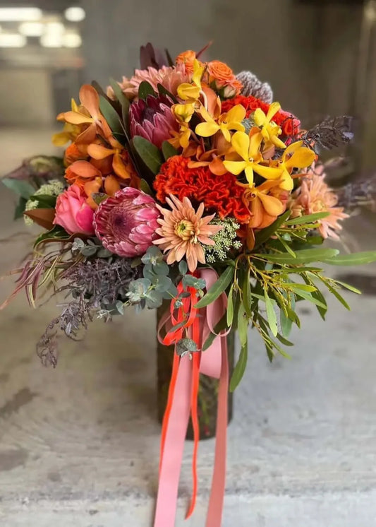 A vibrant autumn floral bouquet featuring orange, pink, yellow, and red flowers with green foliage, tied with a pink ribbon, set against a blurred concrete background.