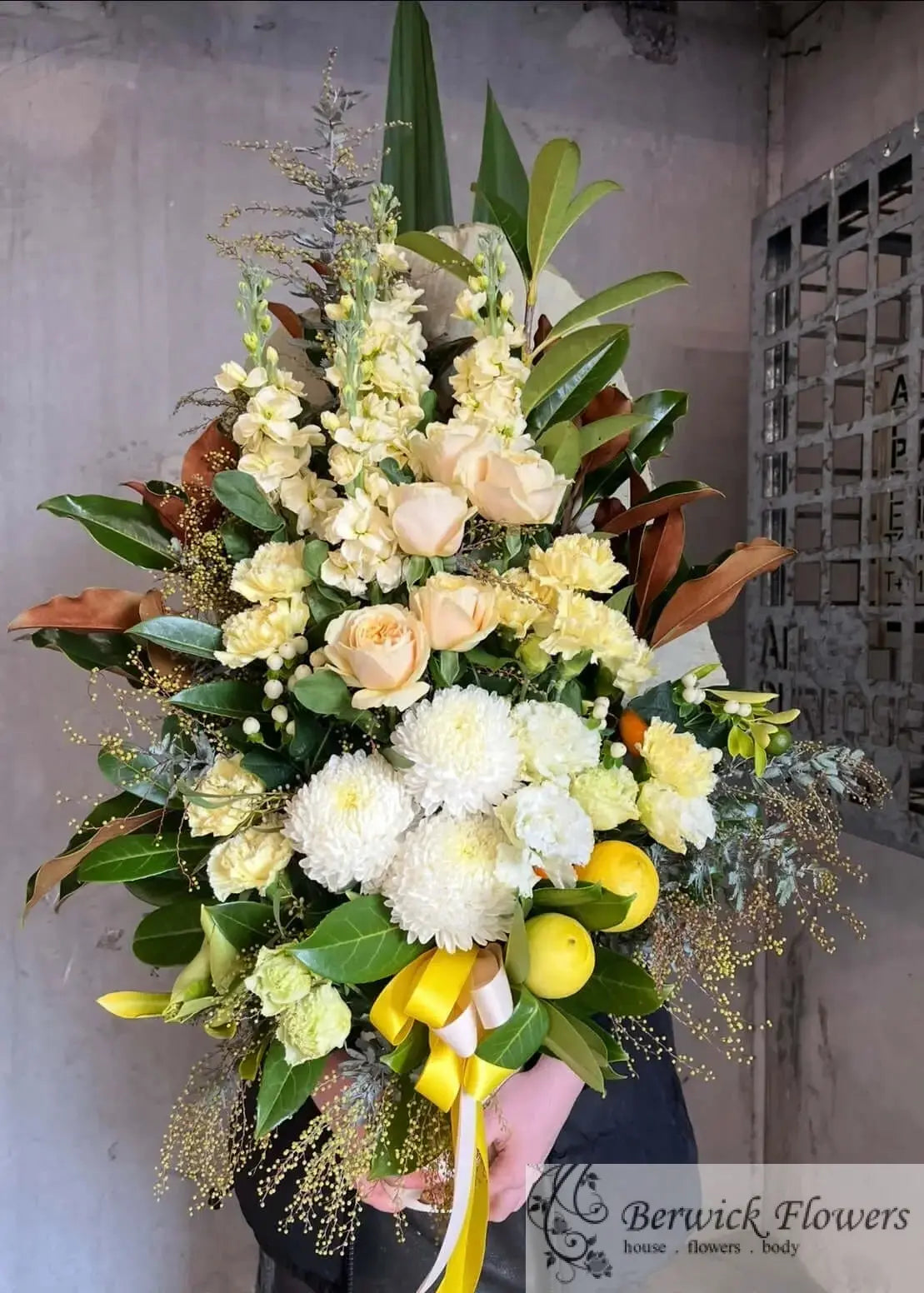 A large, lush floral arrangement held by a person wearing a black jacket. The bouquet features pale yellow roses, white chrysanthemums, green foliage, and bright yellow lemons, tied with a yellow and white ribbon.